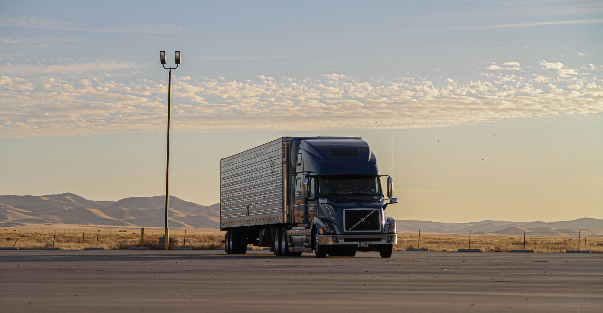 A semi truck is seen on a highway, flanked by impressive mountains in the distance under a sunny sky.